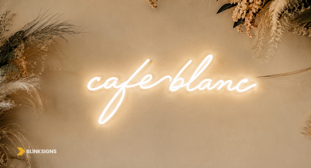 Warm white script neon Cafe Blanc wall sign mounted on a textured boho floral backdrop.